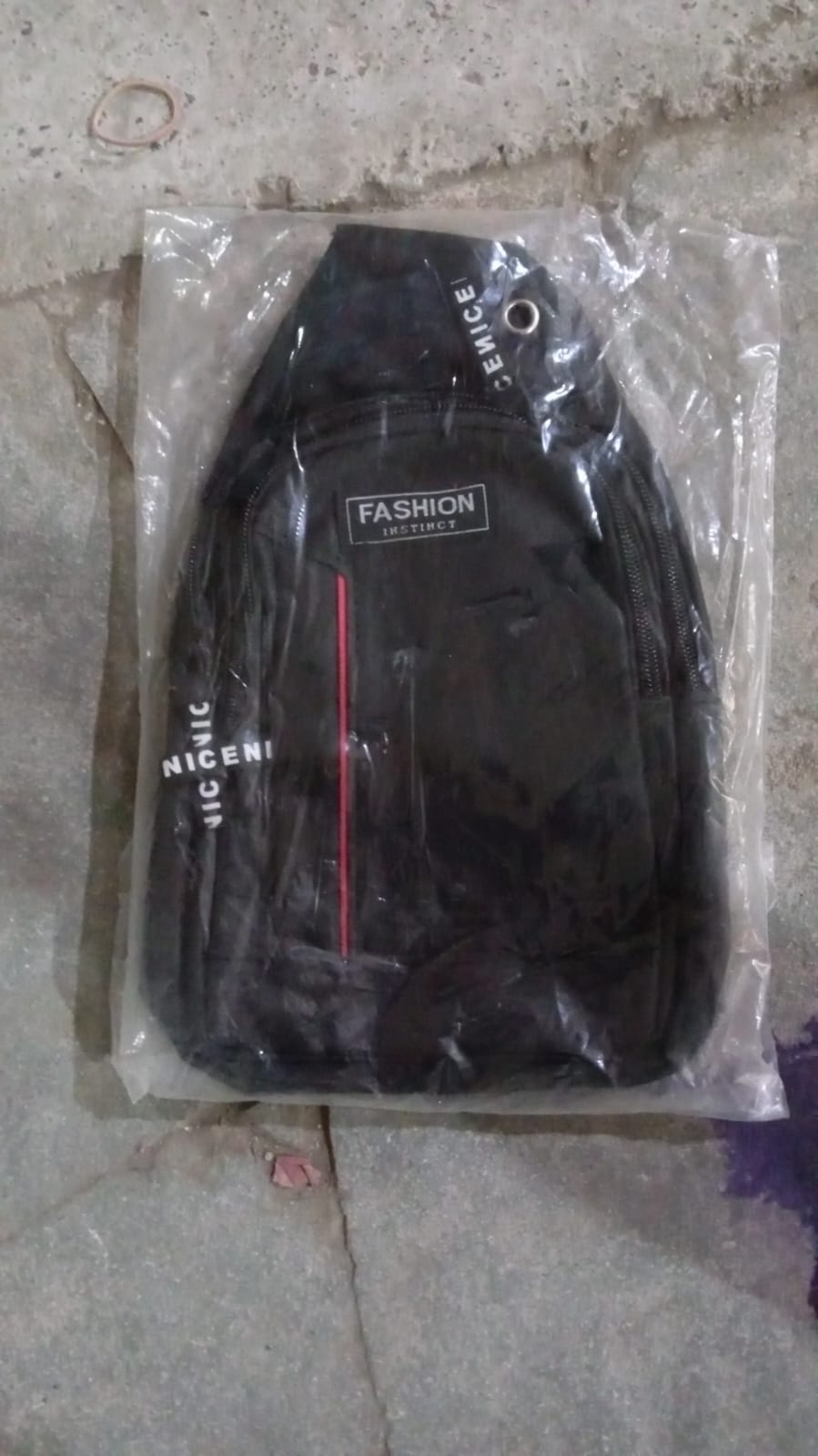 A black sling bag with a red stripe and 'FASHION' label is sealed in clear plastic packaging on a floor.