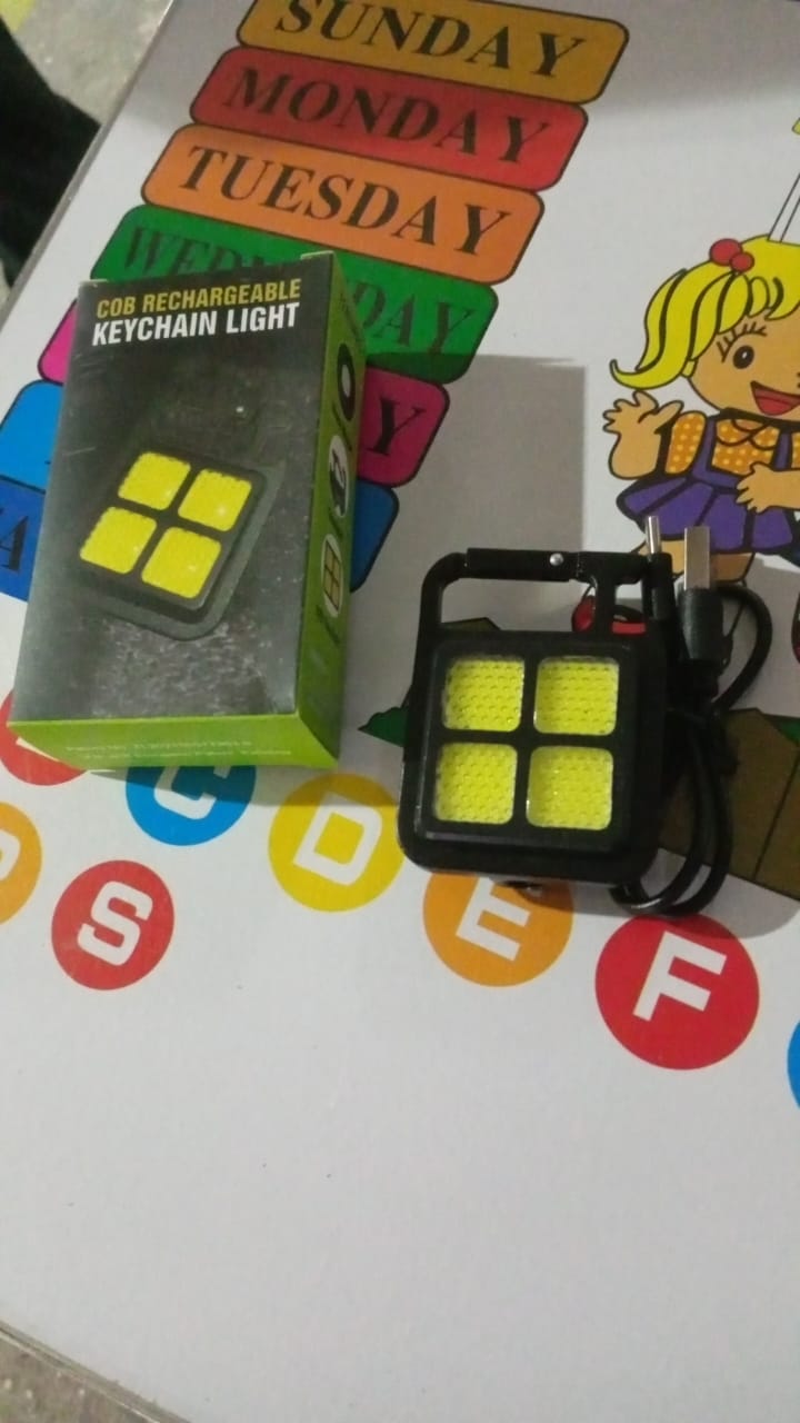 The black LED keychain light with its charging cable and a green product box sit on a white surface with colorful letters