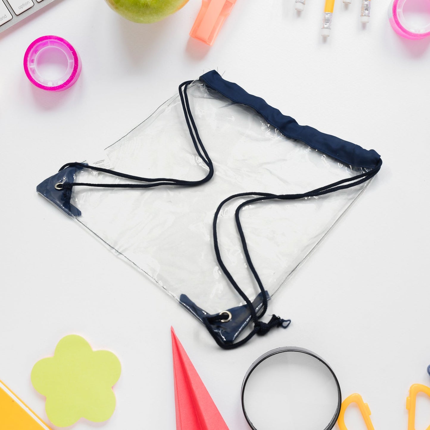 A clear drawstring bag with navy blue accents is centered on a white desk with various office supplies scattered around it.