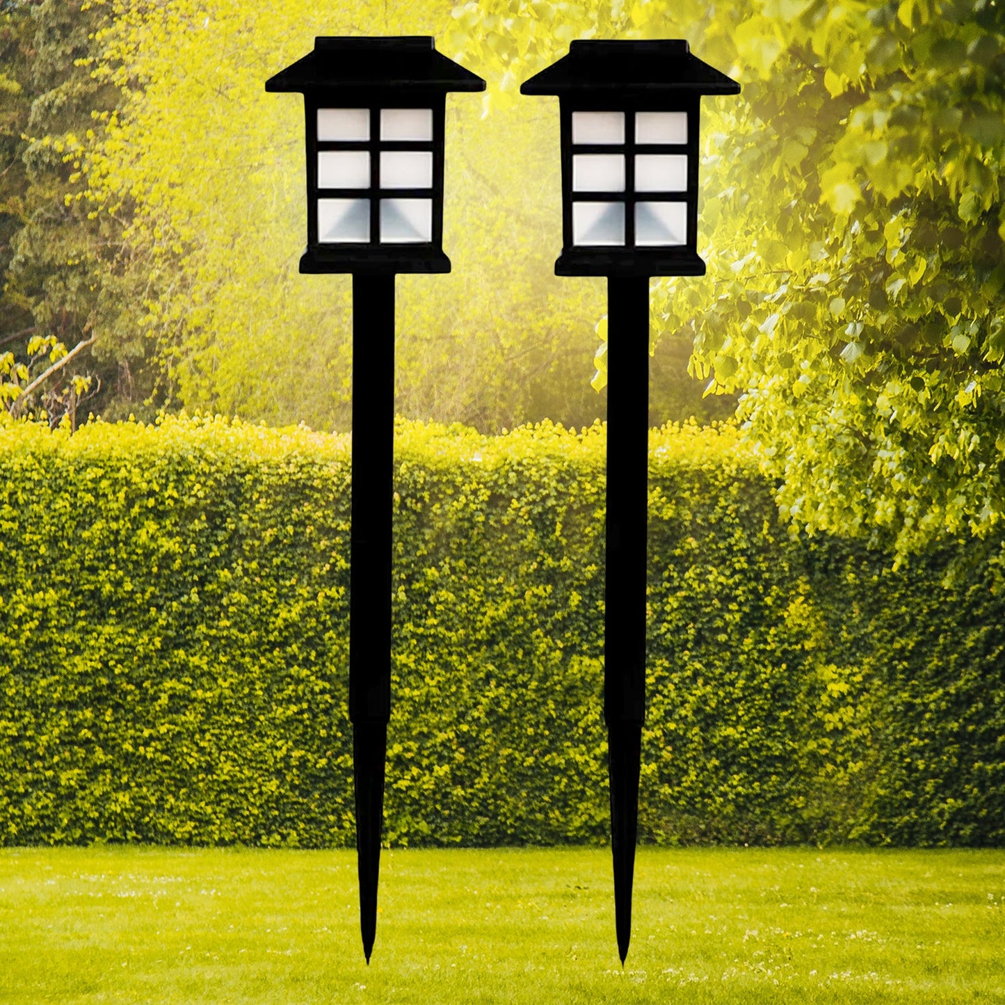 Two black solar garden lights with white paneled tops stand in a vibrant green lawn with hedges and trees.