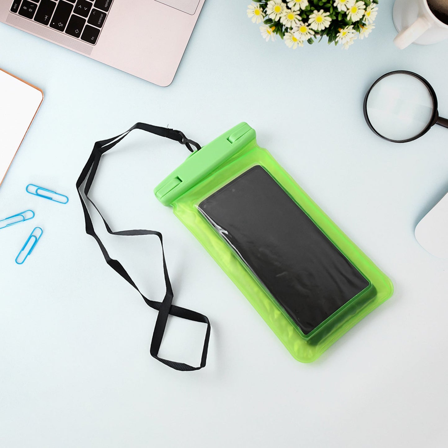 A bright green clear phone pouch with a black lanyard holds a black smartphone on a light blue desk with a silver laptop,