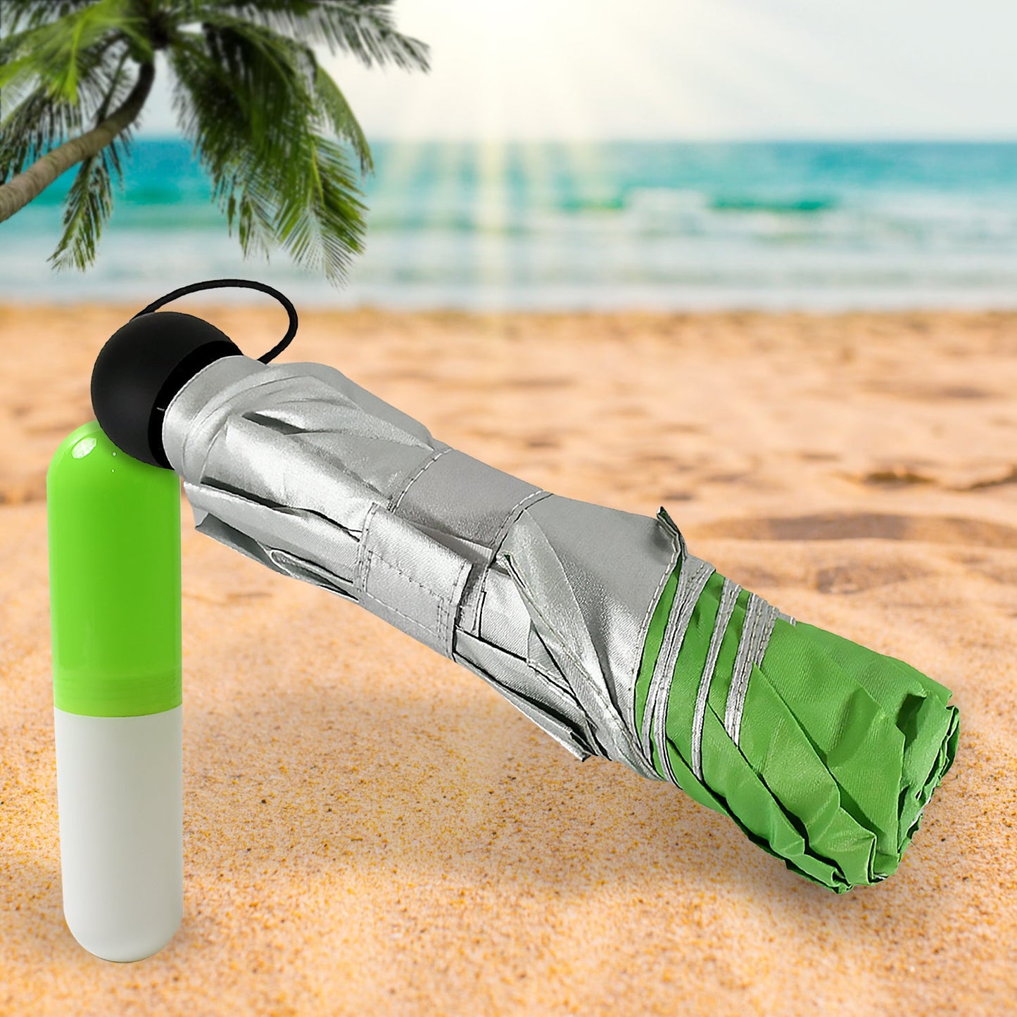 A compact green and silver umbrella is folded next to its capsule case on a sandy beach, with a palm tree and ocean in the