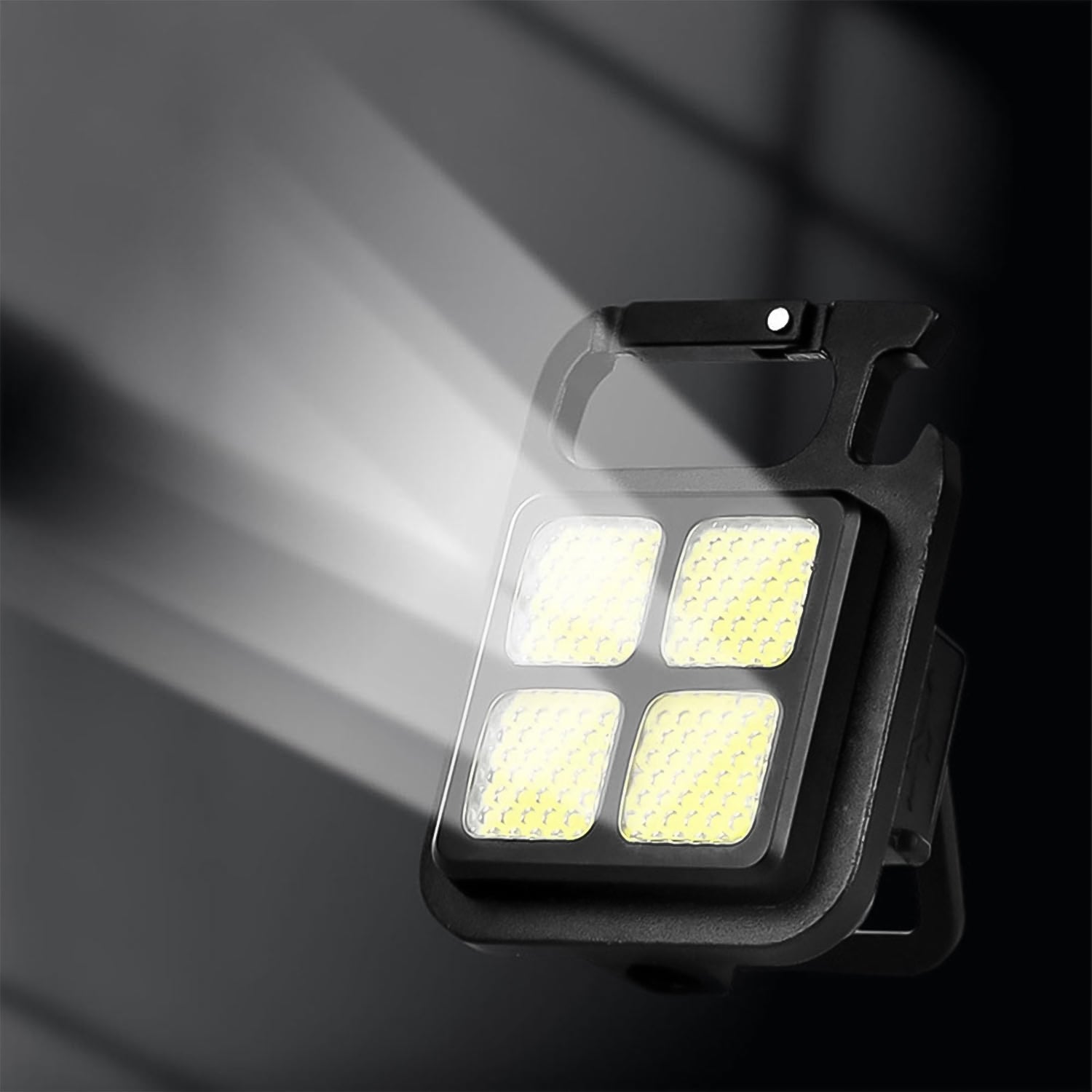 The black LED keychain light emits a strong white beam from its four yellow squares against a dark background.