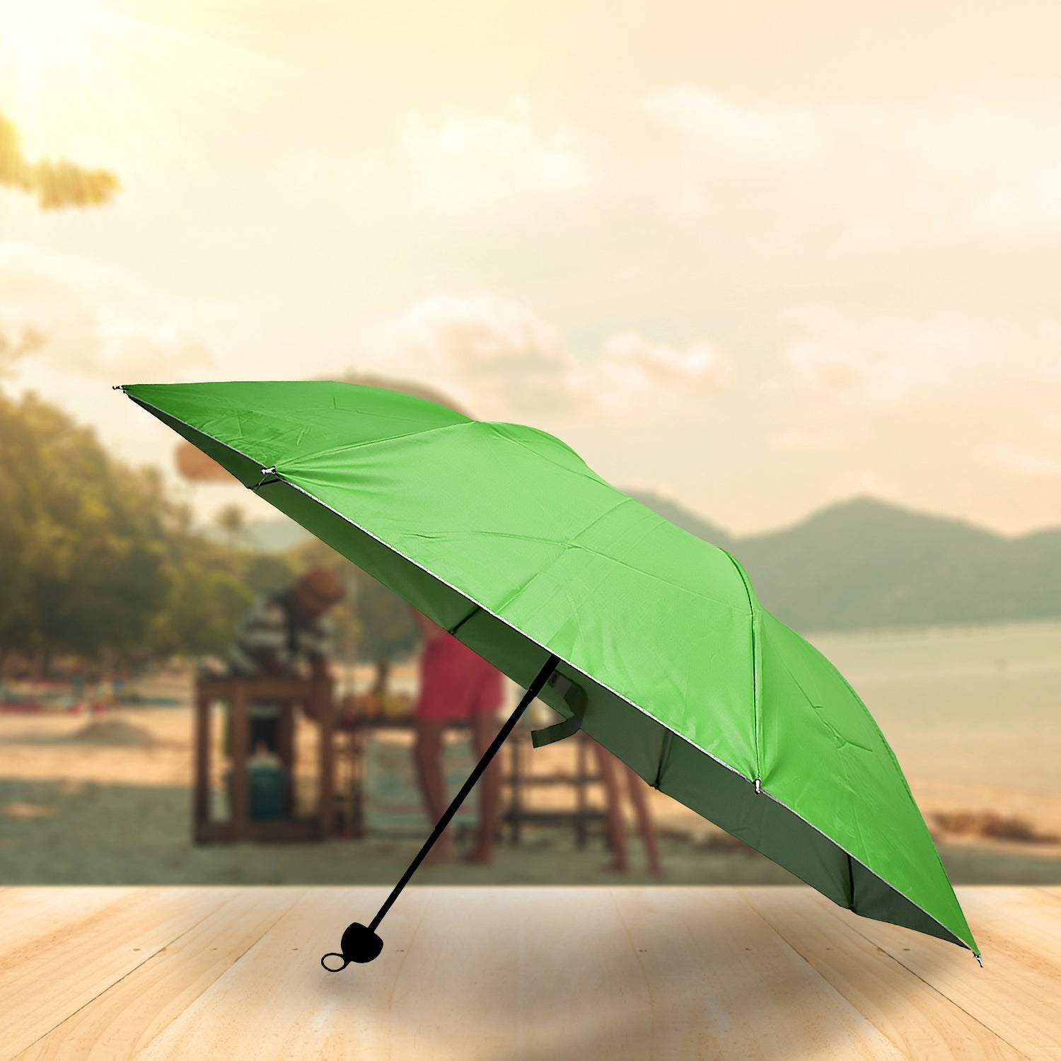 A green umbrella with a black handle is open on a wooden surface, with a blurry beach scene in the background.