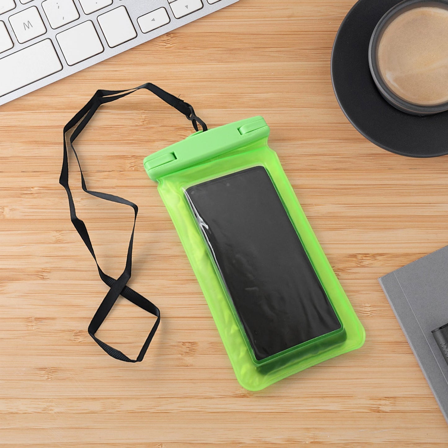 A green clear phone pouch with a black smartphone and lanyard is on a wooden desk next to a white keyboard and black coffee