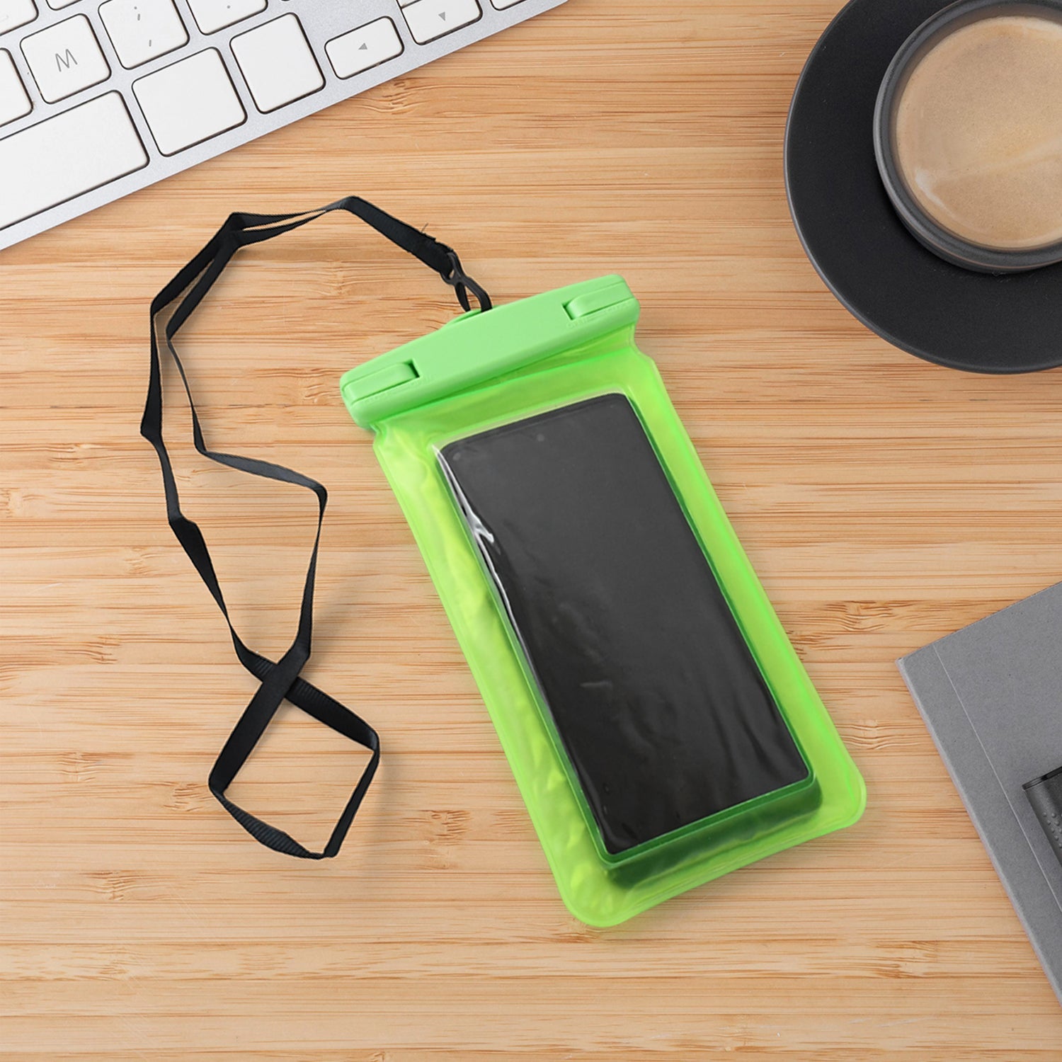 A green clear phone pouch with a black smartphone and lanyard is on a wooden desk next to a white keyboard and black coffee
