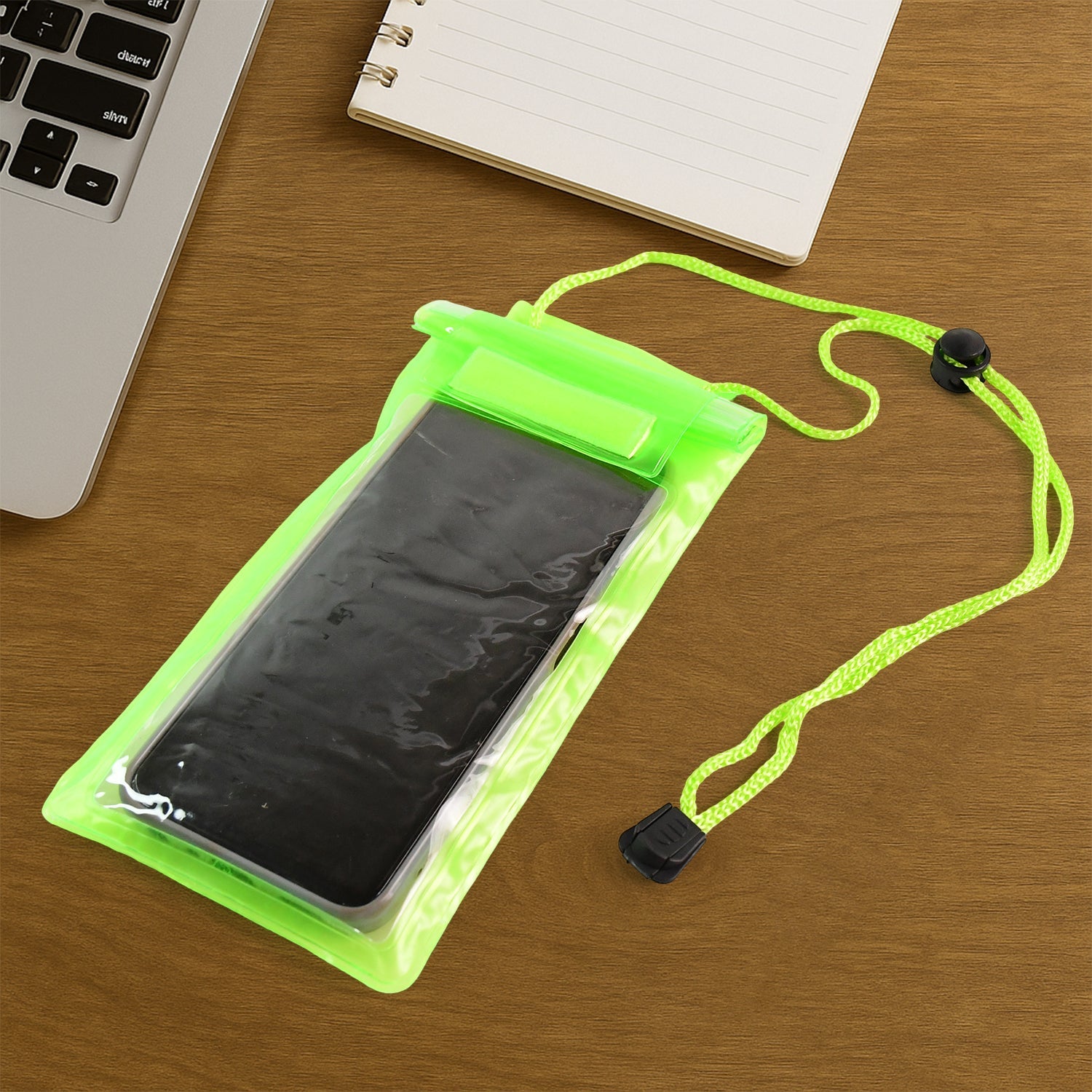 A bright green waterproof phone pouch containing a dark phone lies on a wooden desk next to a laptop and a notebook.