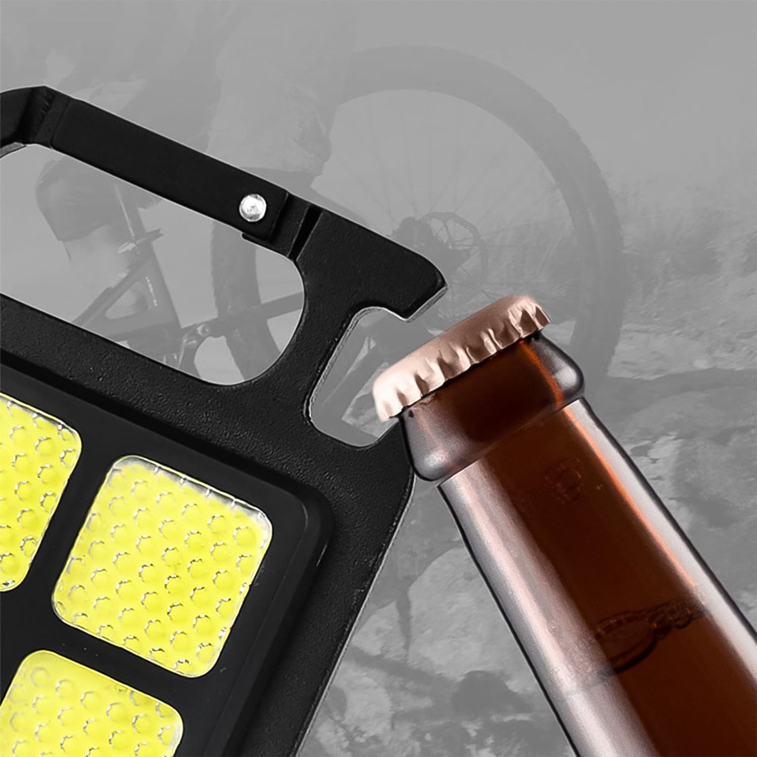 A close-up of the black LED keychain light's bottle opener feature, opening a brown glass bottle, with a blurred bicycle in