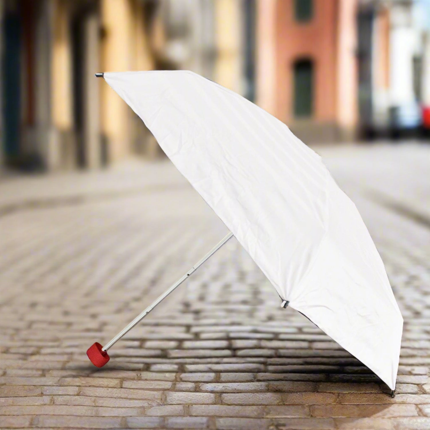 A white umbrella with a red handle stands open on a cobblestone street, ready for use.
