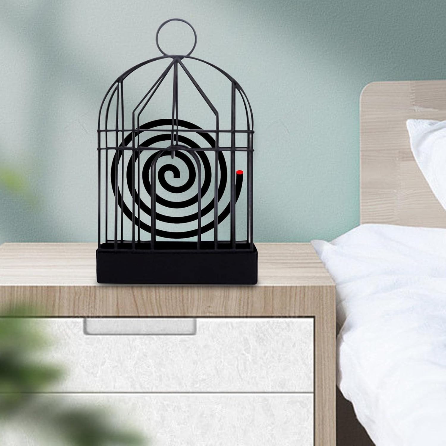 A black iron birdcage-style holder with a burning mosquito coil sits on a light wood nightstand next to a bed.