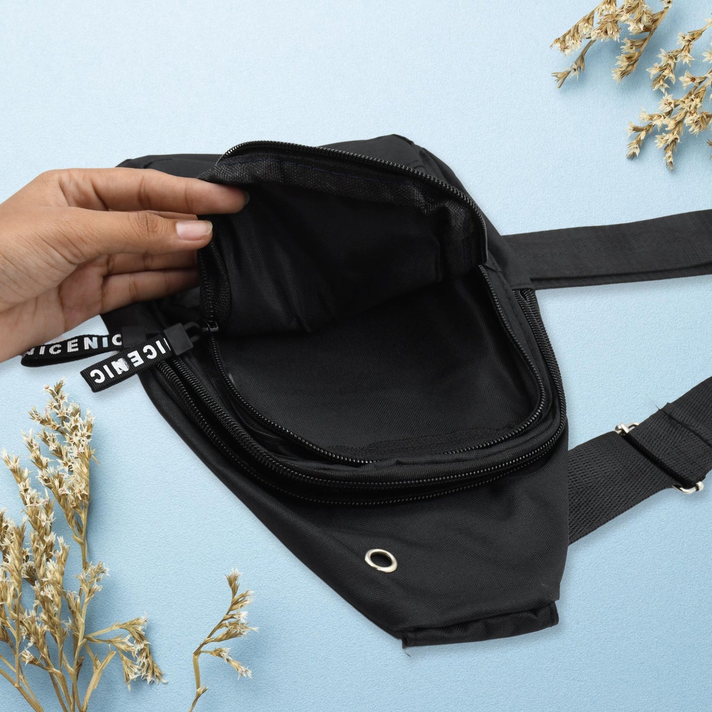 A hand opens the main zipper compartment of a black sling bag, revealing the empty interior.