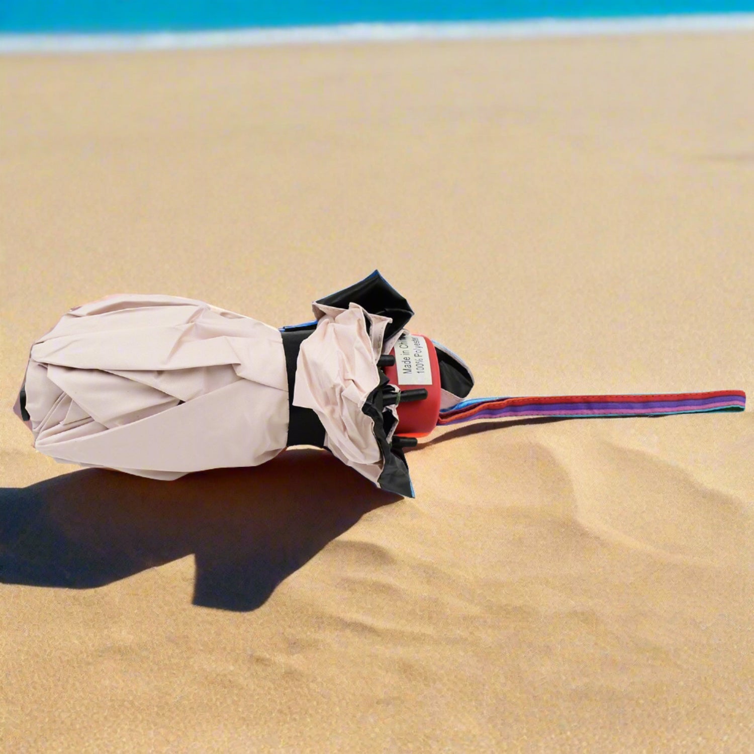 A folded beige umbrella with a red handle and rainbow strap rests on a sandy beach.
