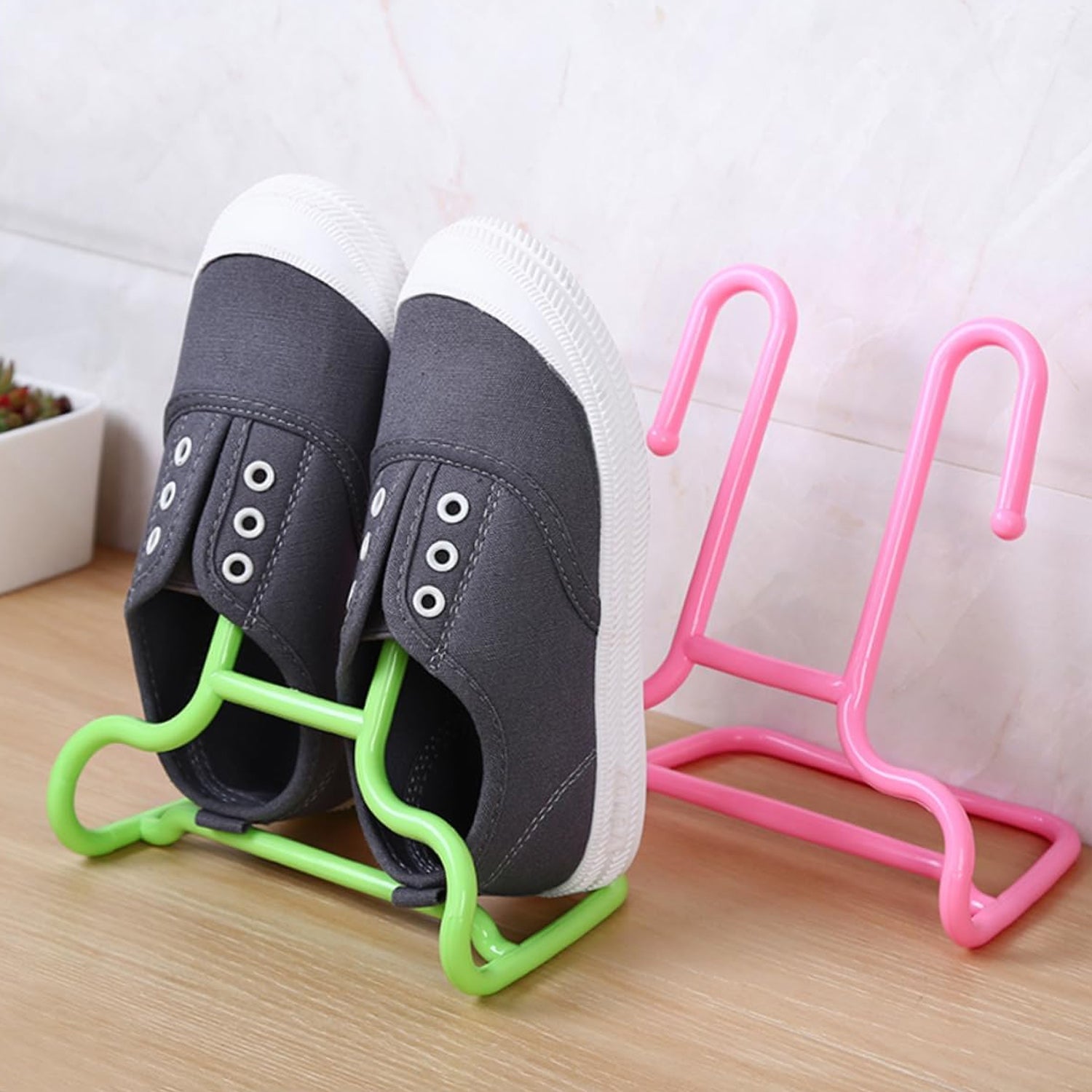 A pair of grey canvas shoes hangs from a green plastic shoe drying rack, with a pink rack standing nearby on a wooden
