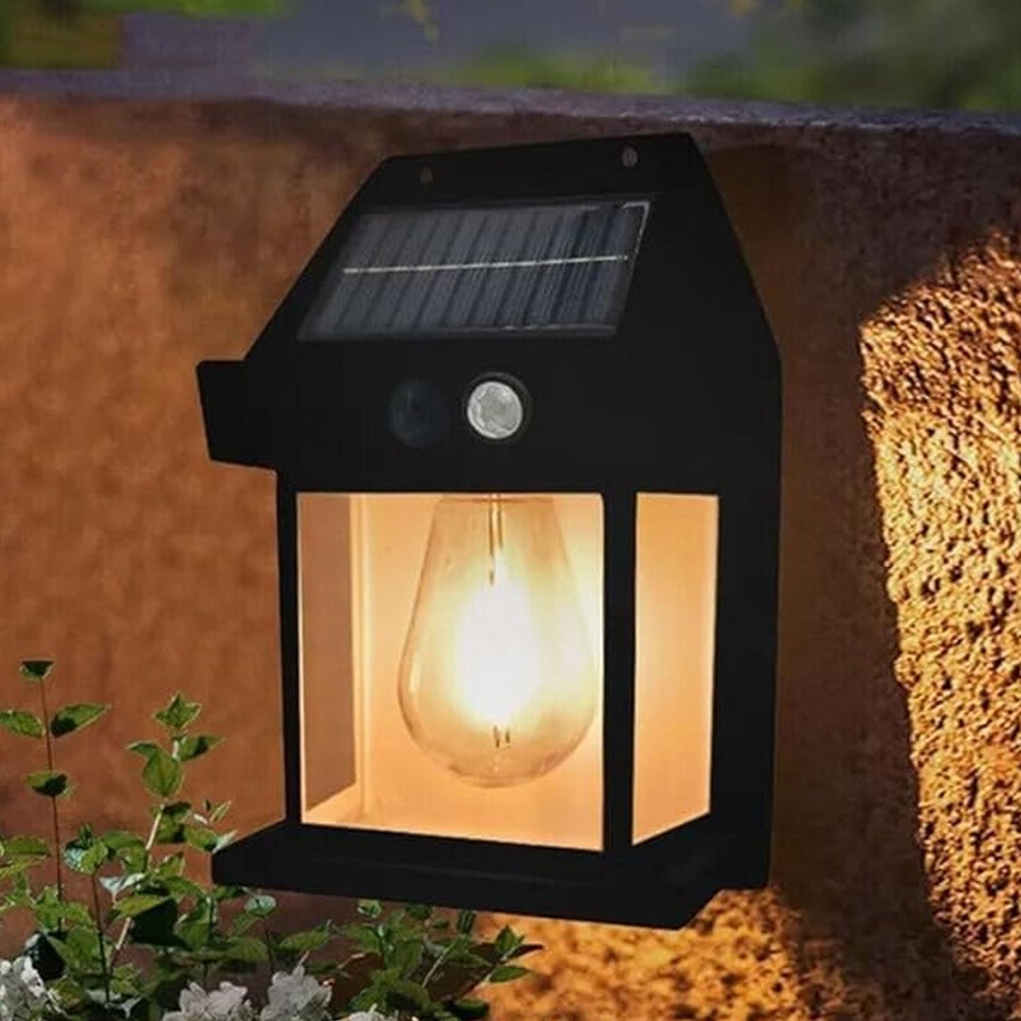 A black solar wall lamp with a clear bulb glowing warmly is mounted on a textured brown wall above green plants.