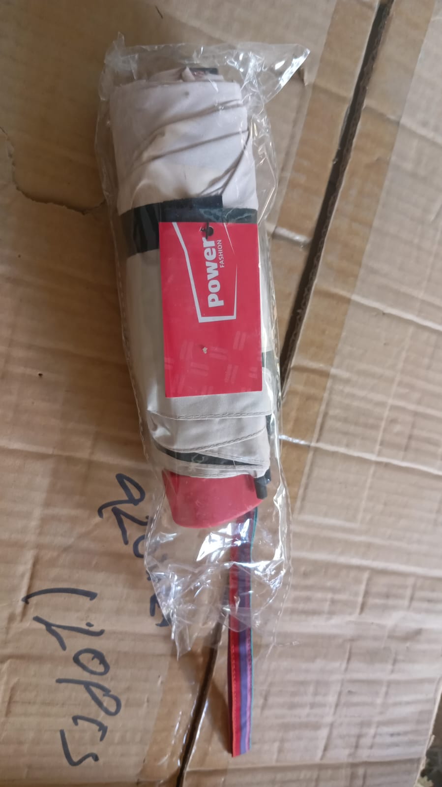 A folded beige umbrella with a red handle and rainbow strap is sealed in clear plastic packaging.