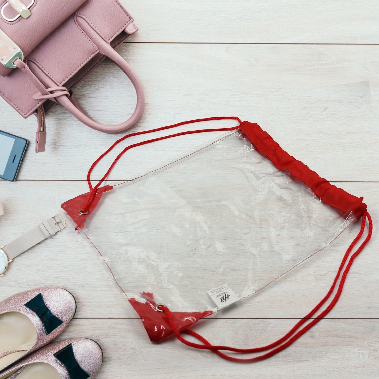 A clear ClassiCarry drawstring bag with red accents lies on a white wooden floor surrounded by a pink purse, phone, watch,