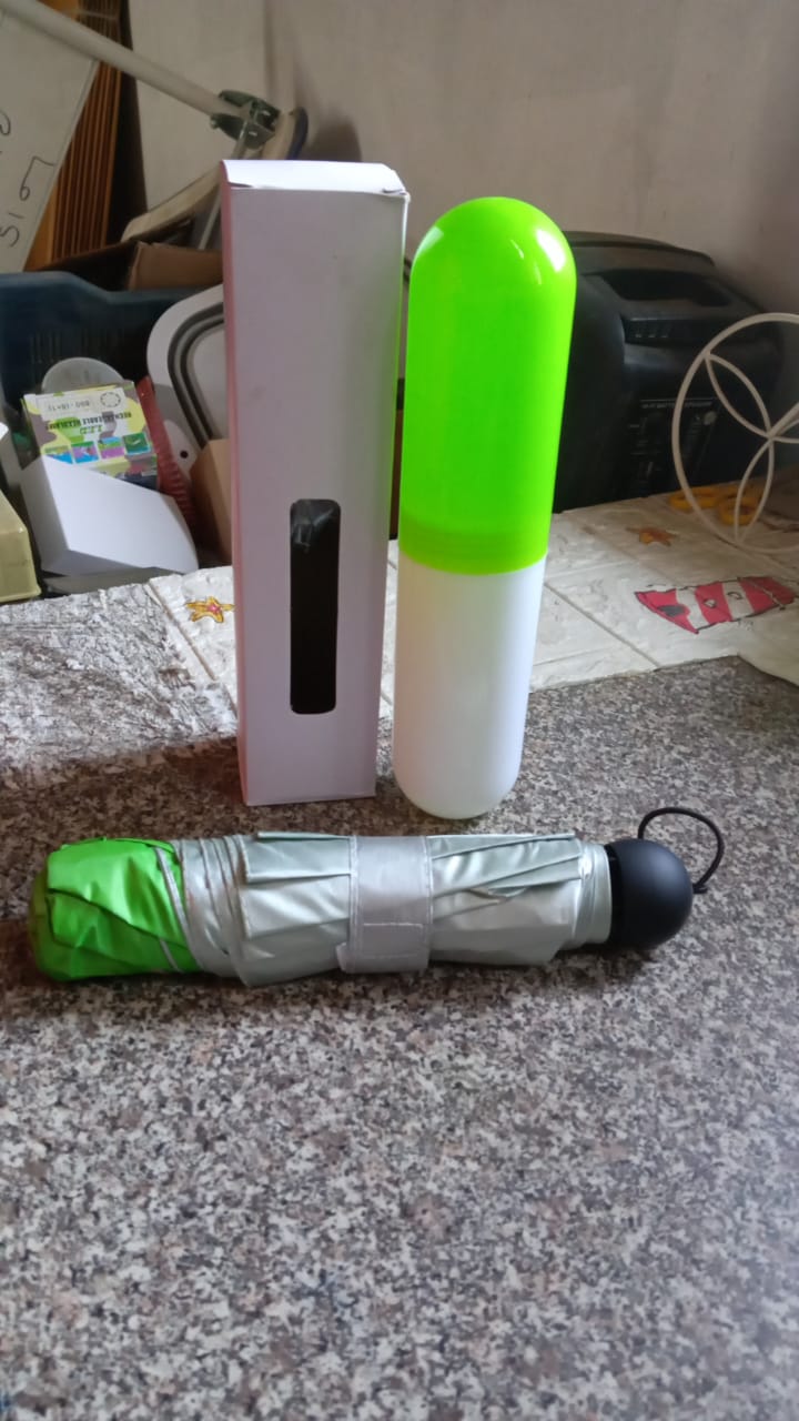 A folded green and silver umbrella lies on a speckled floor next to its white box and a green and white capsule case.