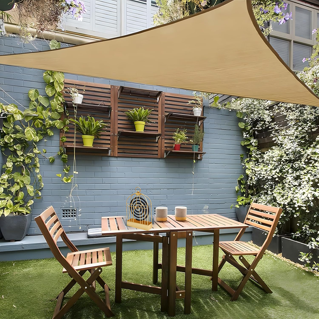 A gold birdcage-style mosquito coil holder sits on a wooden table in a lush green outdoor patio setting.