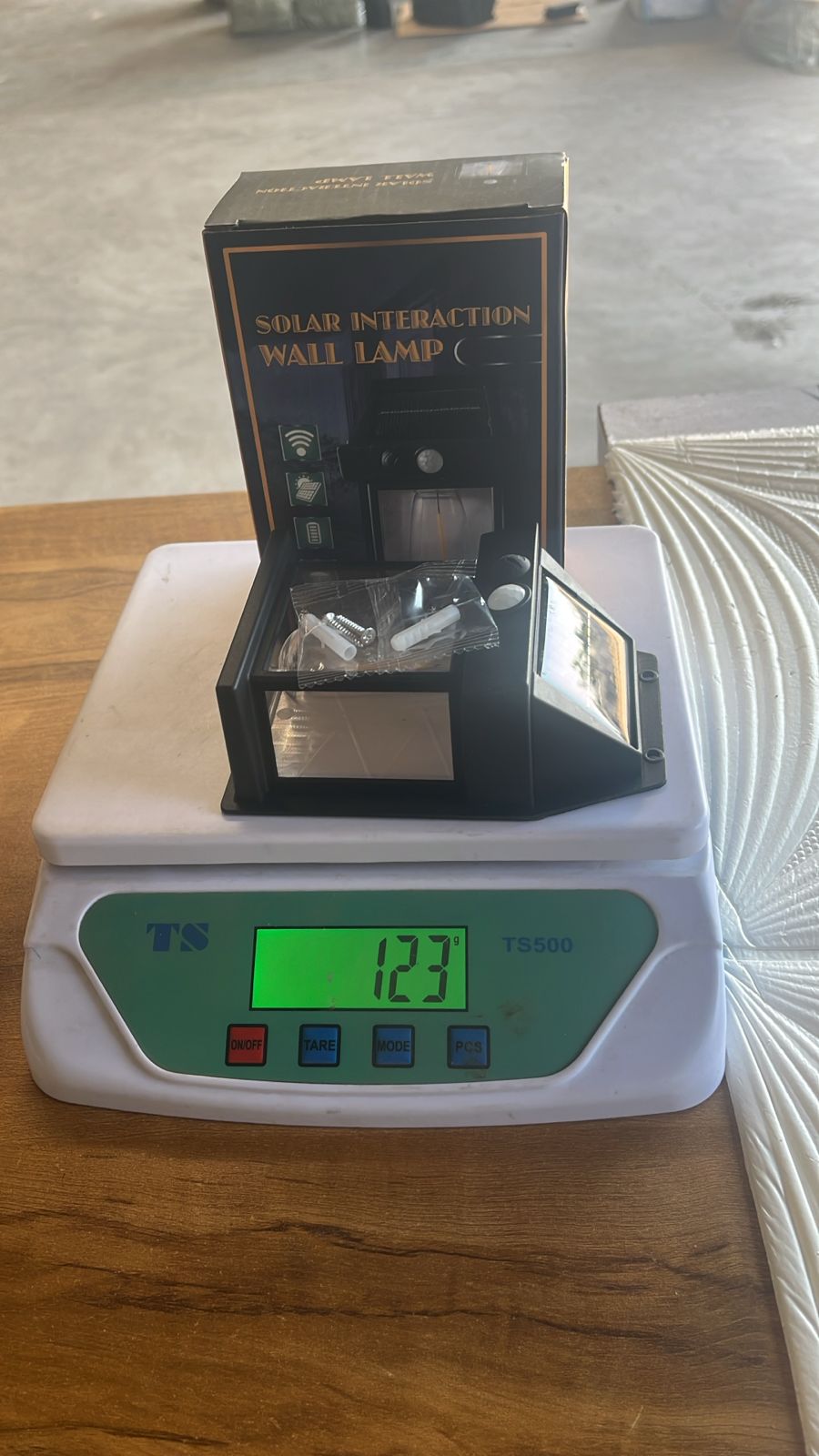 A black solar lamp and its open box, including mounting hardware, are being weighed on a white digital scale displaying 123.
