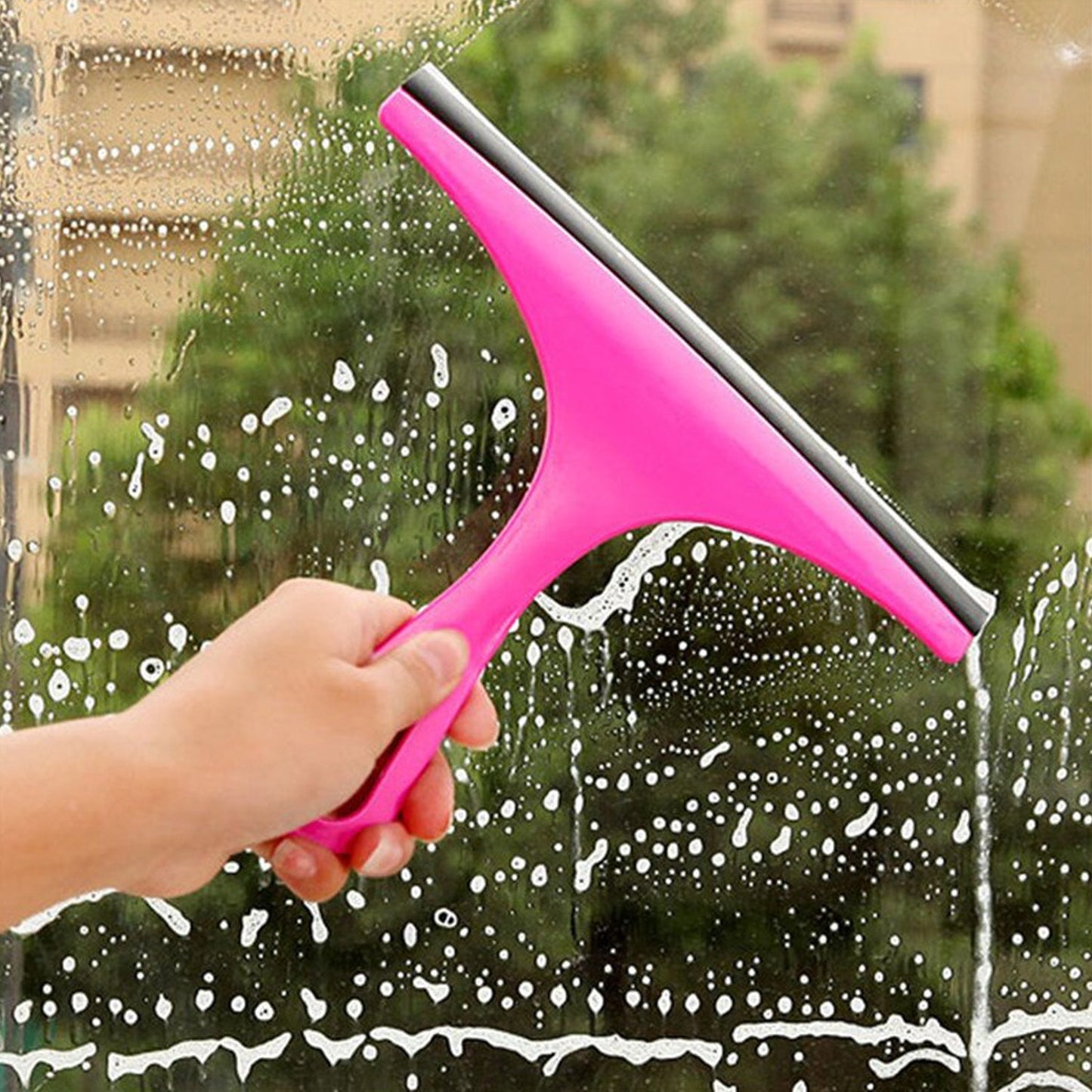 A hand holds a pink car mirror wiper, cleaning a window covered in white suds with a green background.