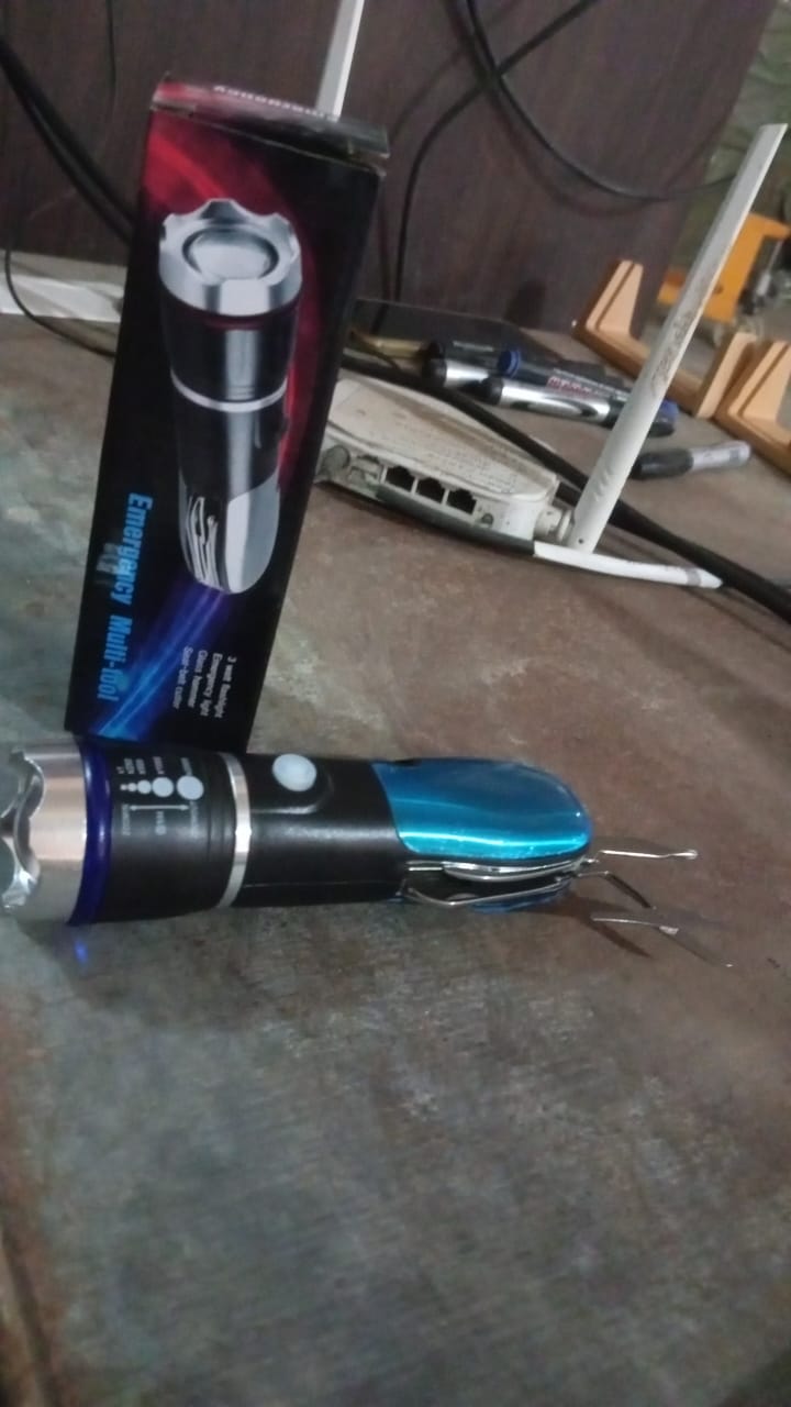 A black and blue multi-tool LED flashlight lies on a table next to its black and red retail box, with electronics in the