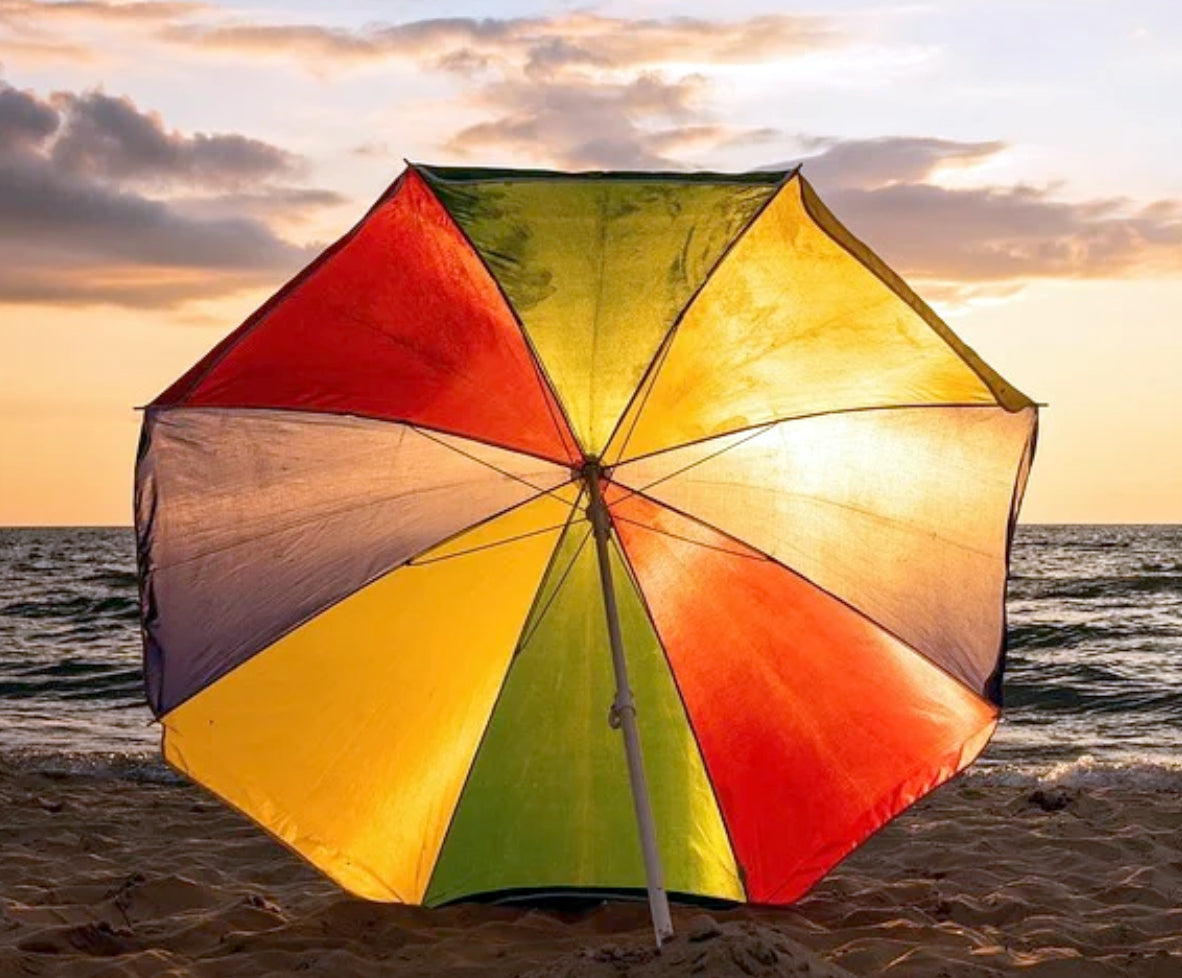 A colorful DrizzlePro umbrella with red, yellow, green, and blue panels stands on a beach at sunset, with the sun low over