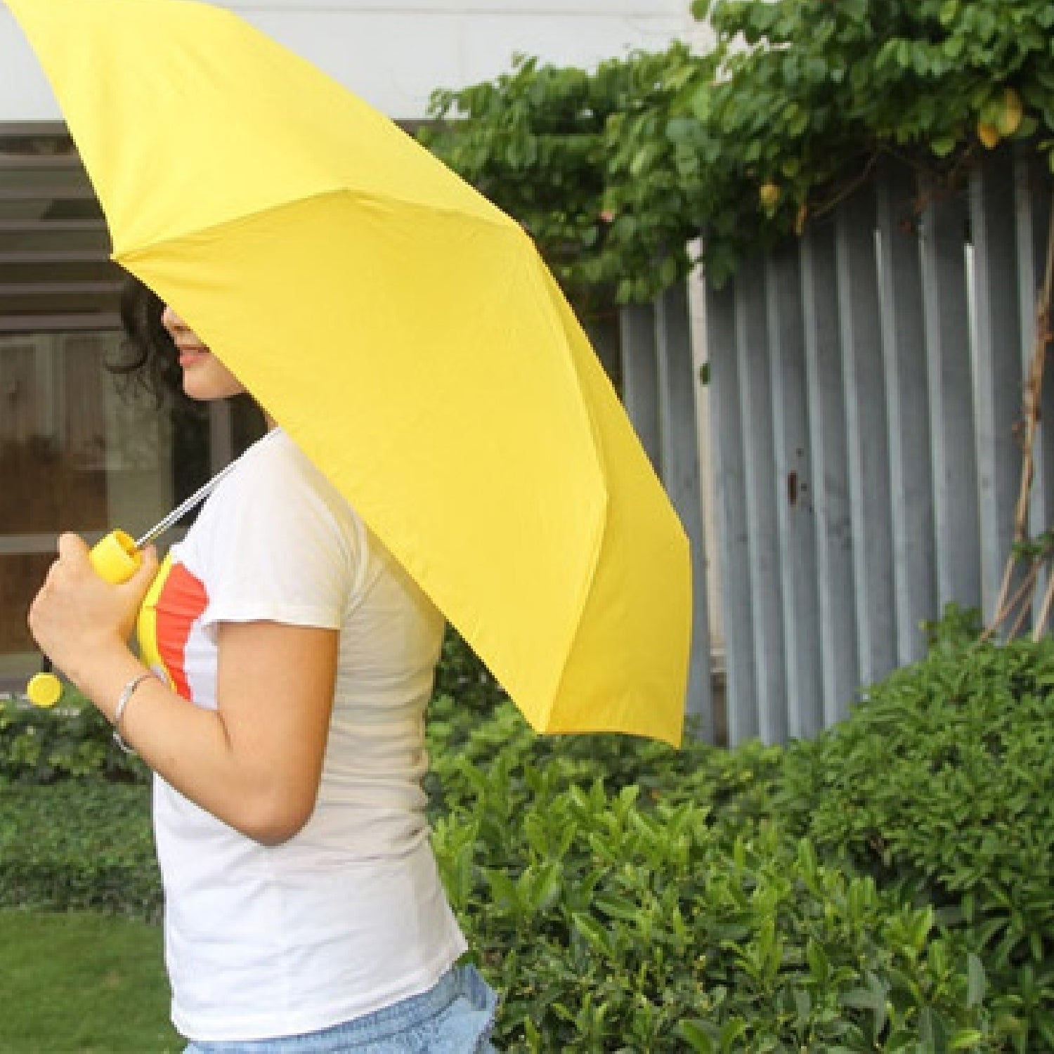A person from the waist up holds an open yellow umbrella, walking past green bushes and a fence.