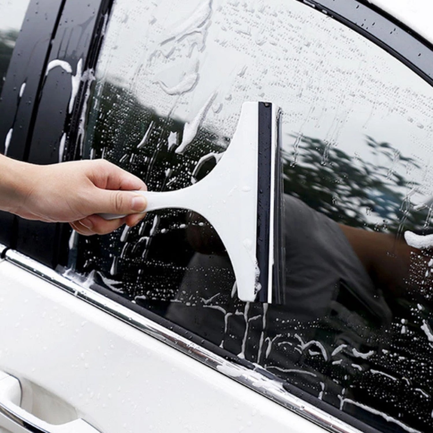 A hand uses a white car mirror wiper to clean a car window, leaving streaks in the white suds.