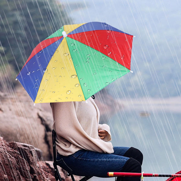 A person in a beige sweater and jeans sits by a lake, wearing a colorful red, yellow, green, and blue Umbrella Hat in the