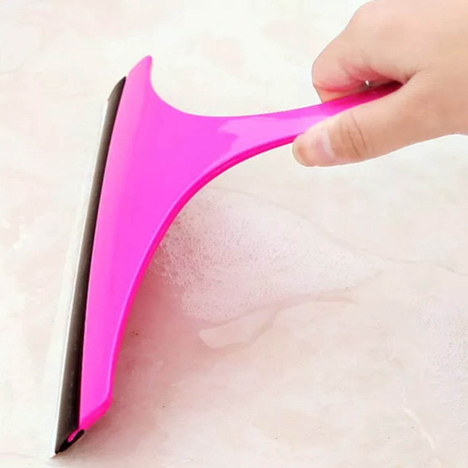 A hand holds a pink car mirror wiper, wiping suds from a light-colored tiled surface.