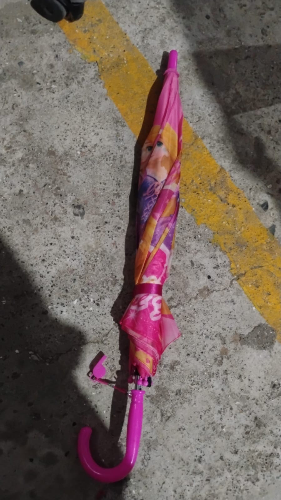 A closed pink umbrella with princess designs lies on a concrete ground with a yellow painted line.