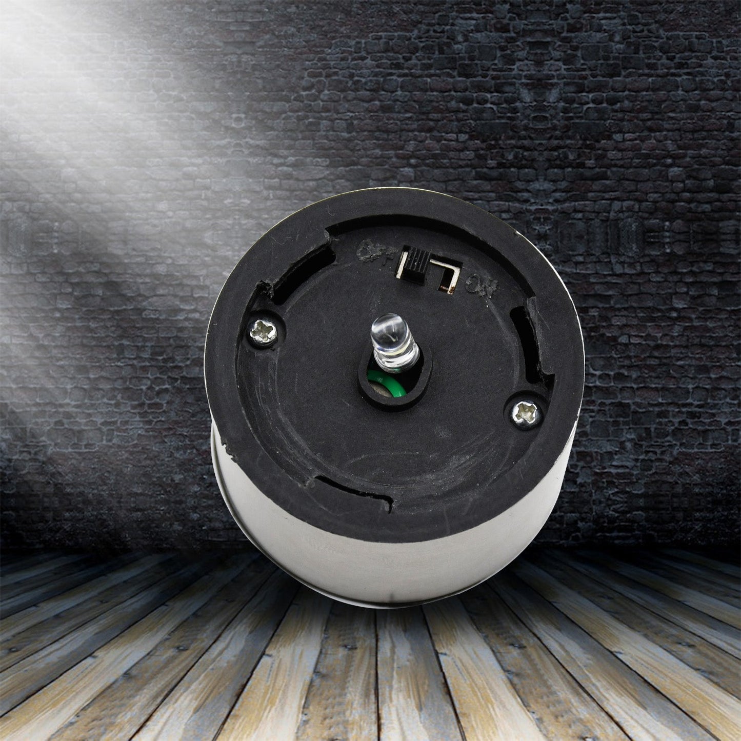 The round silver and black solar cap with an LED bulb is positioned against a dark brick wall and wooden floor.