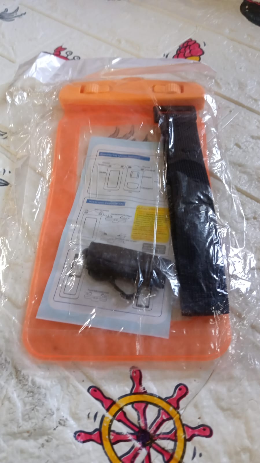 An orange waterproof phone pouch, black lanyard, and instructions are sealed in clear plastic packaging on a patterned