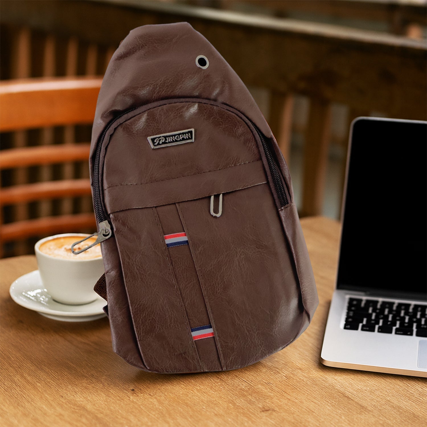 A brown leather sling bag rests on a wooden table next to a white coffee cup and a silver laptop.