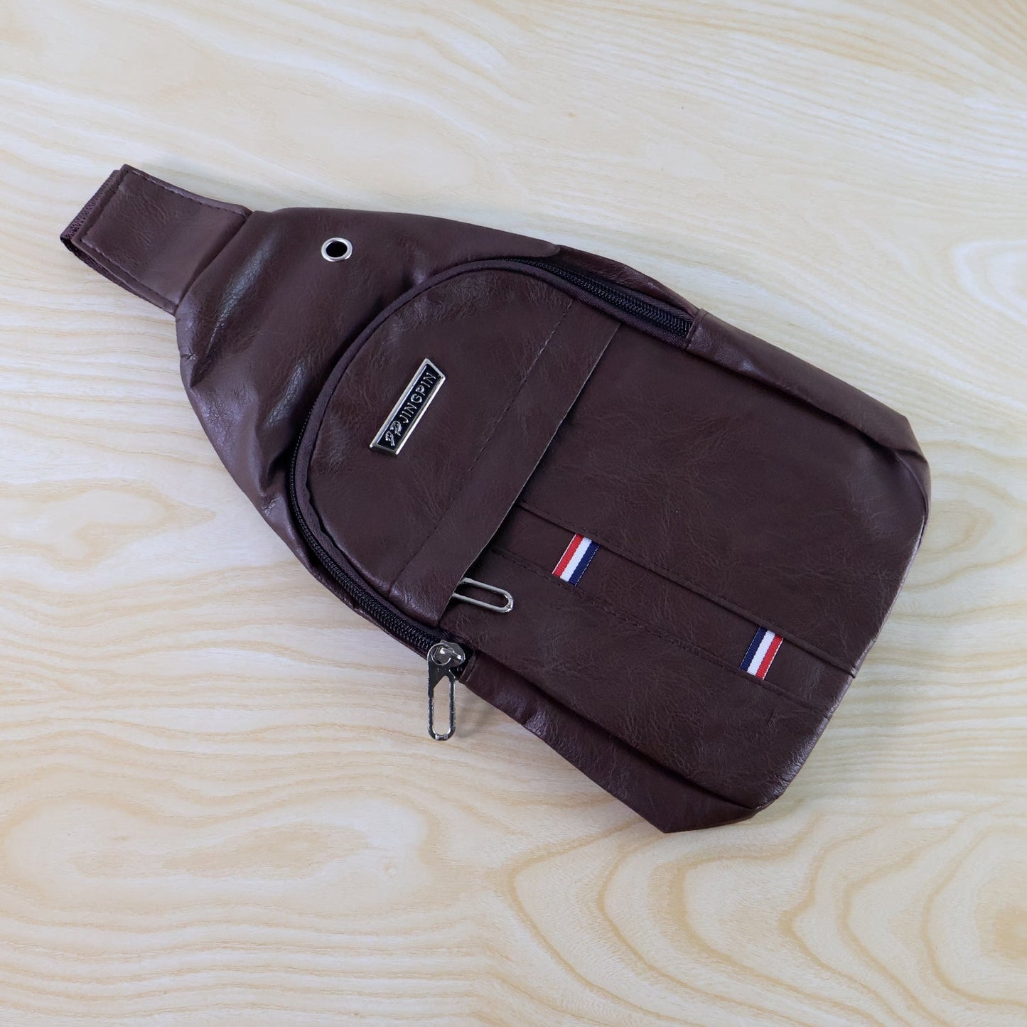 A brown leather sling bag with a textured surface and a prominent silver zipper pull on a wooden background.