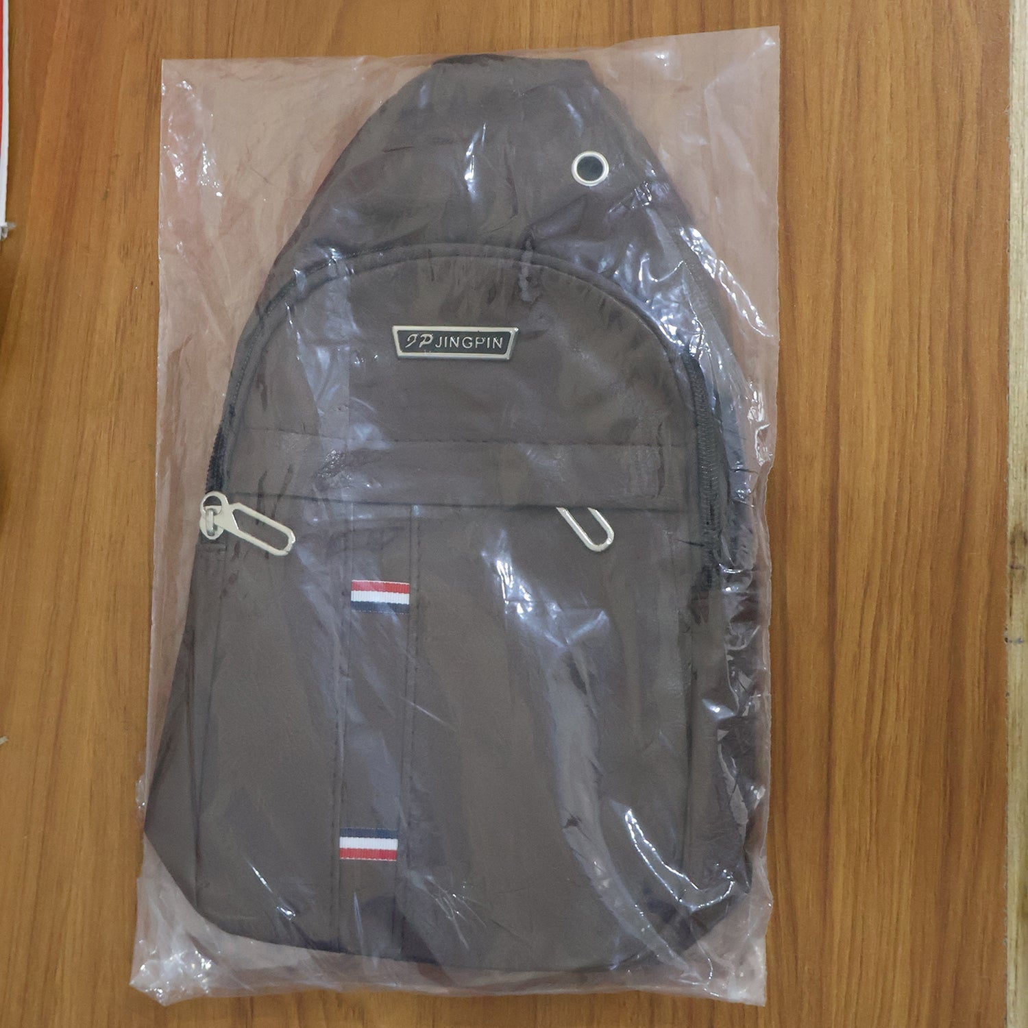 A brown leather sling bag is sealed inside a clear plastic bag, showcasing its front pockets and strap.