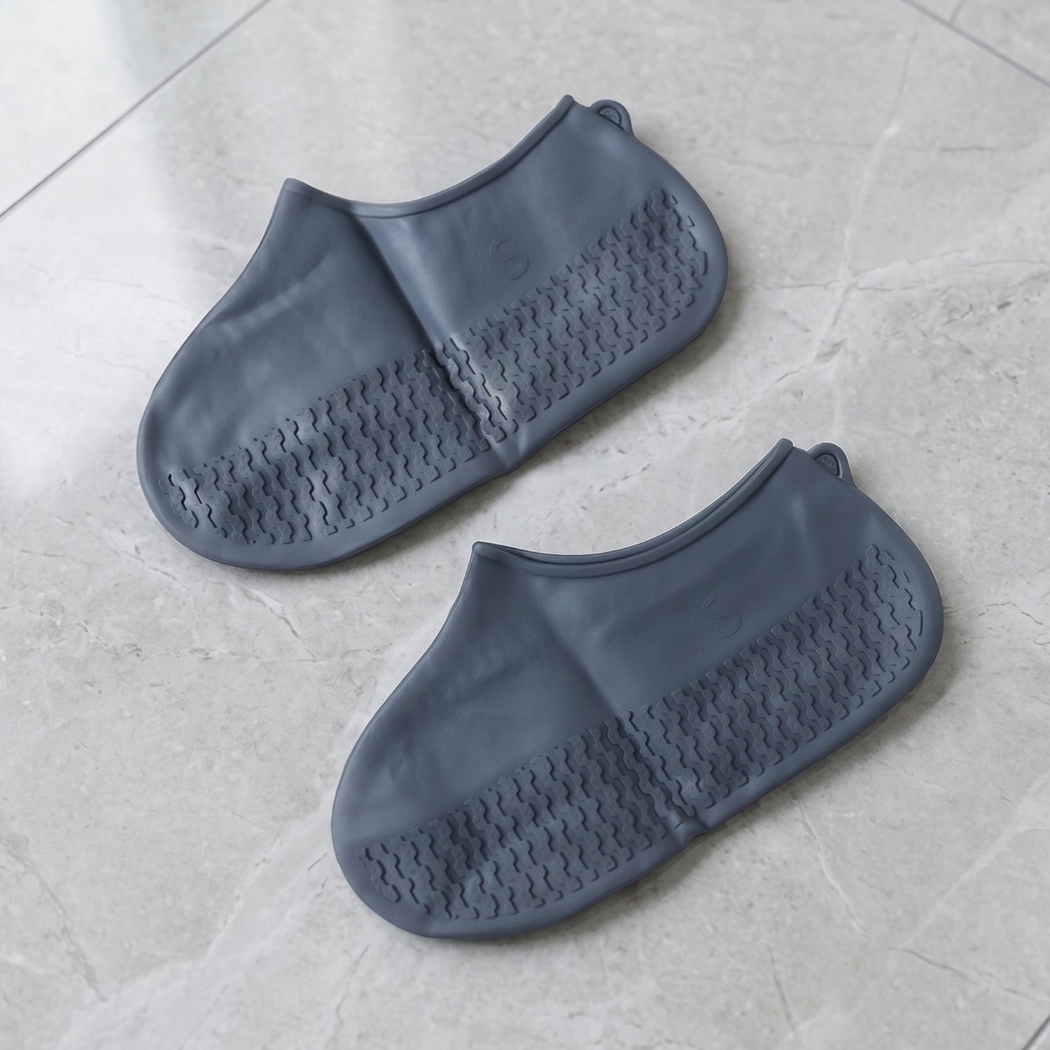 A pair of gray silicone foot coverings with textured soles rests on a light gray tiled surface.