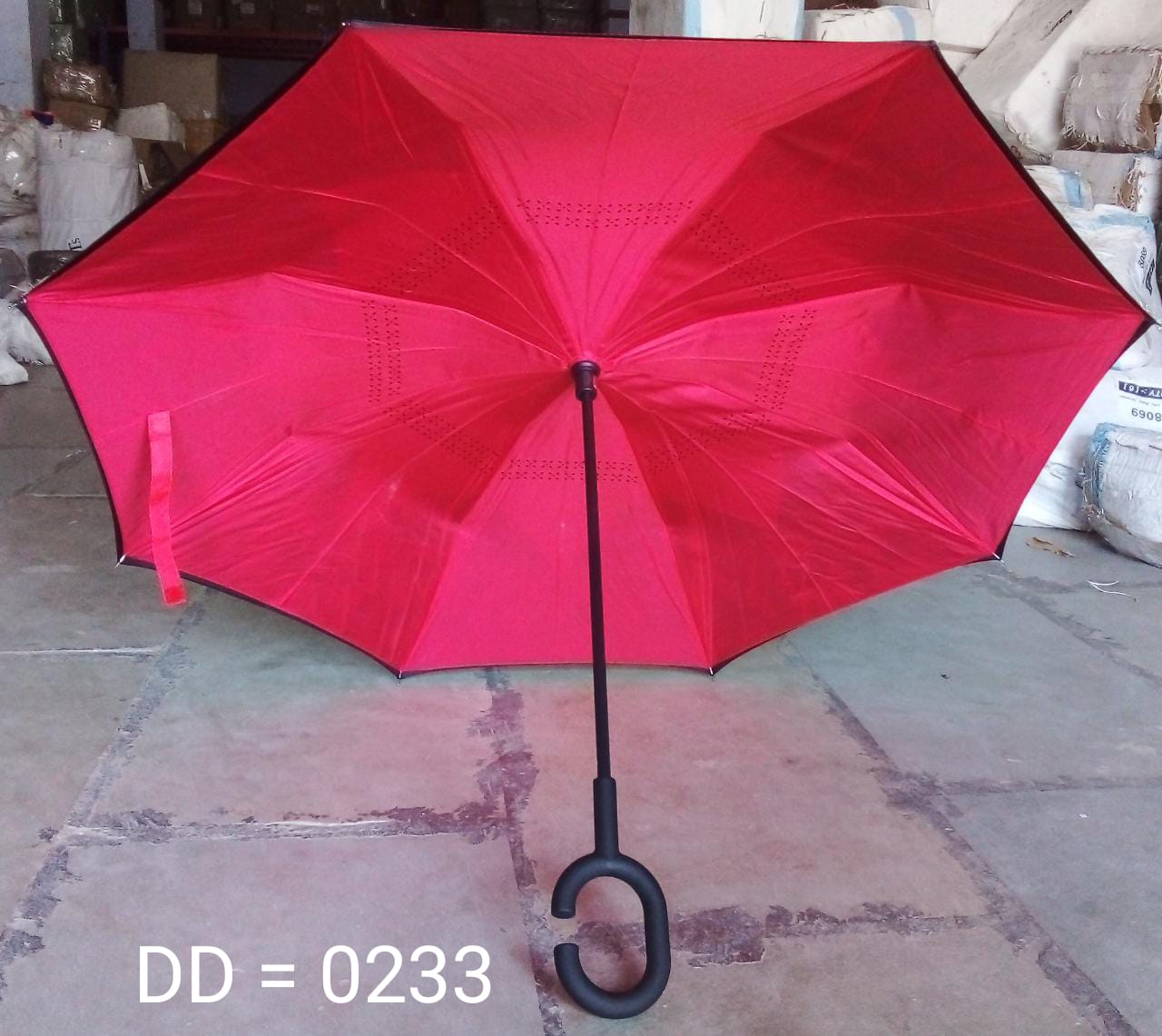 An open, bright red reverse umbrella with a C-shaped handle is displayed on a concrete floor in a warehouse setting.