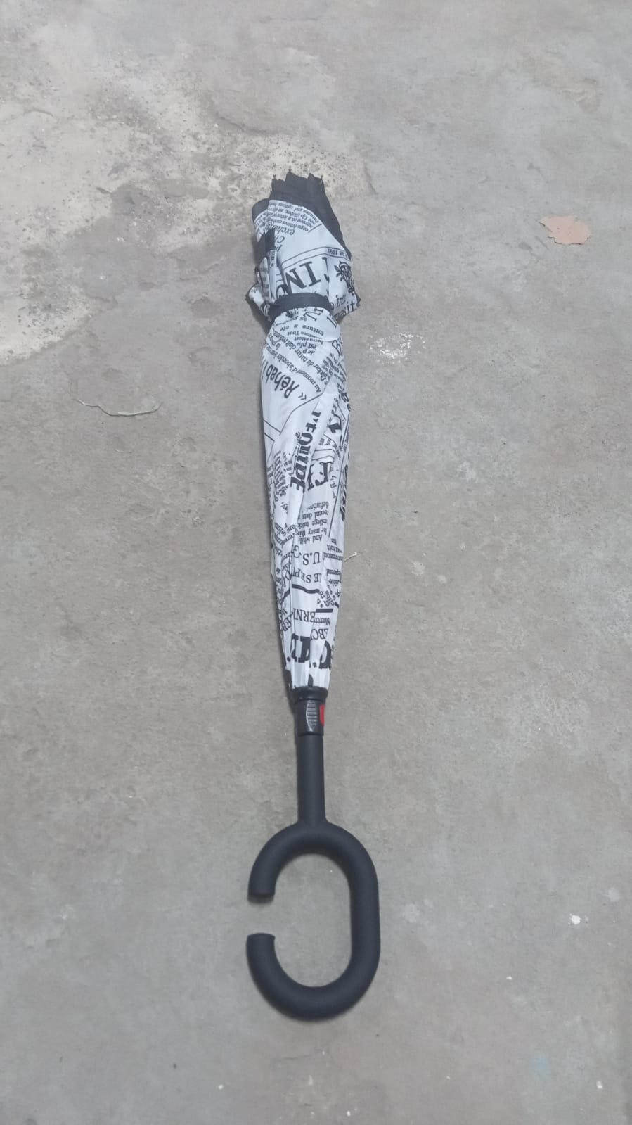 A closed reverse umbrella with a black and white newspaper print is standing upright on a grey concrete surface.