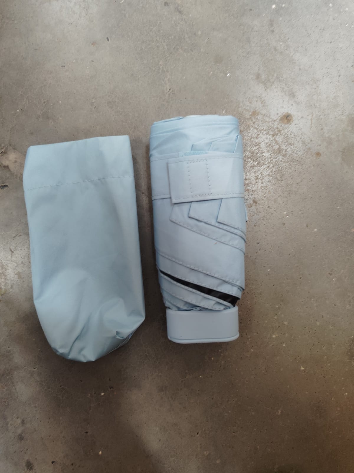 A light blue folded umbrella with a matching storage pouch lies on a concrete surface.